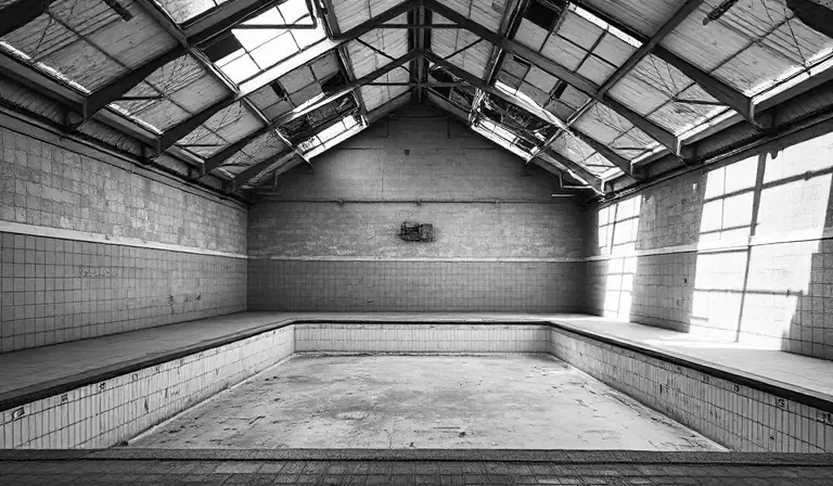 Piscine abandonnée dont le bassin vide se couvre de débris et de végétation.
