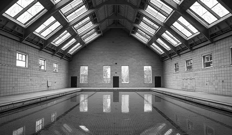 Ancienne piscine intérieure, vidée et dégradée sous une verrière brisée.