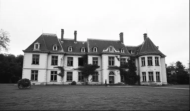 Château abandonné entouré d’un jardin envahi, avec une aile effondrée et des façades couvertes de lierre.